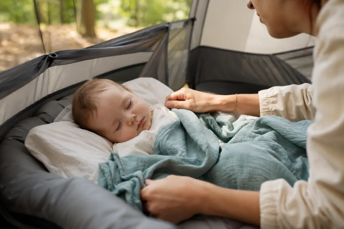 Partir en camping avec bébé : mon guide par âge pour que ça se passe bien Gros plan sur un coin couchage aménagé dans une tente avec un lit parapluie compact et une gigoteuse de bébé